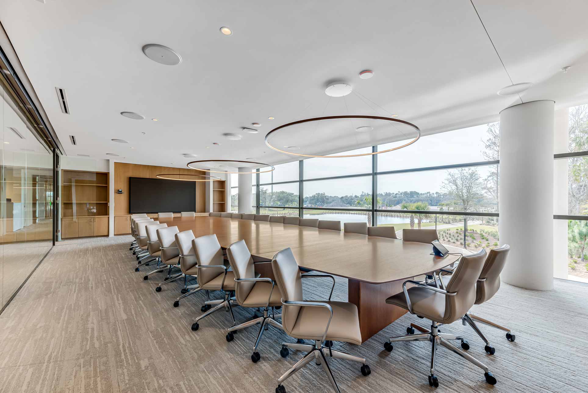 A large wooden conference room with 28 leather rolling chairs lining it. Daylight and Occupancy sensors are visible in the ceiling and the lush landscaping of the grounds are visible through the glass exterior wall. Hanging over the table are statement circular pendants.