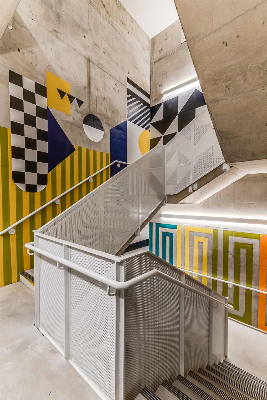 A winding industrial-looking staircase with poured concrete steps and walls. A large geometric mural of black & white checkerboard, green and yellow stripes, and others give the space a virbrant feel.