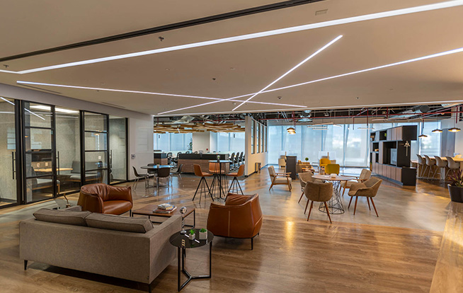 A landscape view of Sekura's Offices. Dramatic linear light criss-crosses the ceiling to form an abstract pattern. Visible are a seating are with leather chairs, private meeting rooms with glass doors, and open office area. 