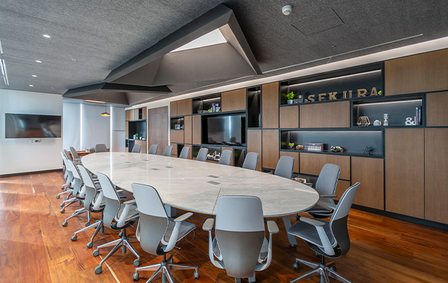 One of Sekura's conference rooms. a large stone oval table dominates the room, with 20 gray plastic rolling chairs lining it. Integrated bookcases line the room and a geometric skylight is in the ceiling.