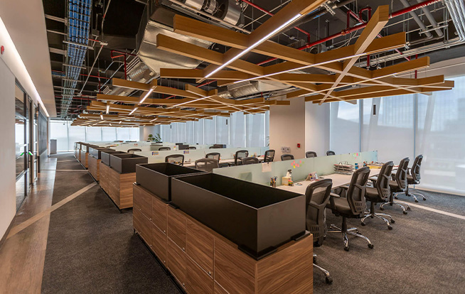 A statement geometric wooden pendant with integrated linear lighting  hangs below an exposed ceiling. Employee desks sit in rows to create an airy, open workspace